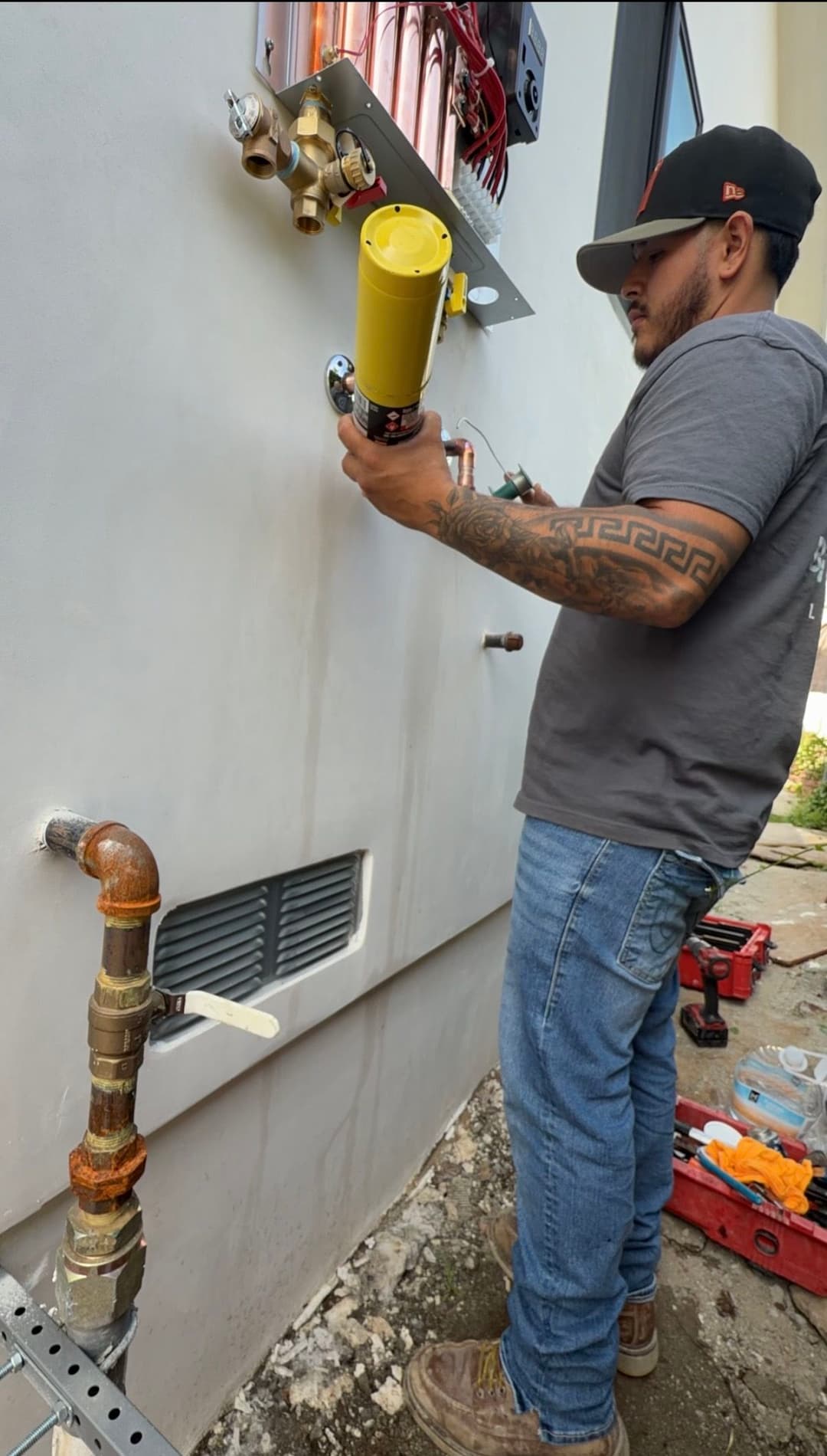 Plumber installing equipment on exterior wall, tools and valves visible.