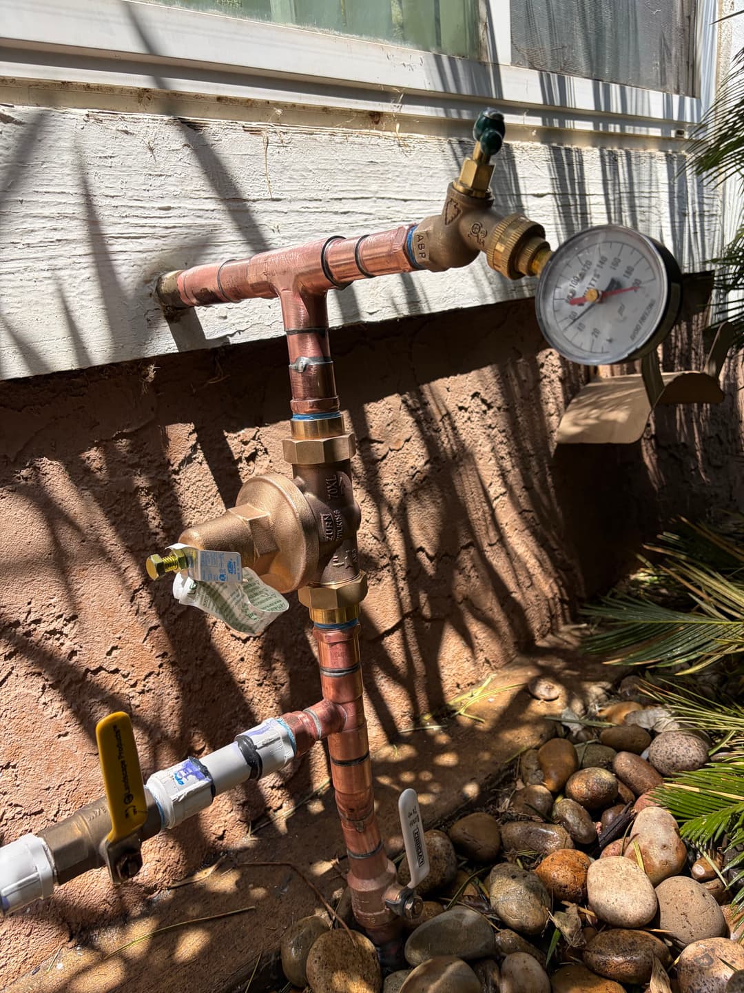 Water pressure gauge and valve on copper plumbing outside a house, surrounded by rocks.