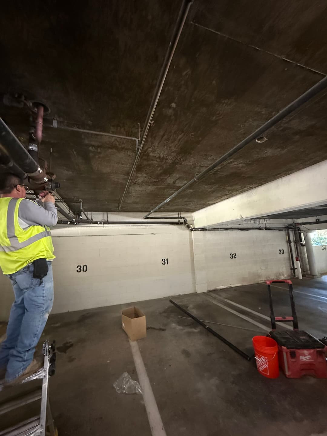 Worker repairing pipes in a commercial parking garage with marked parking spots.