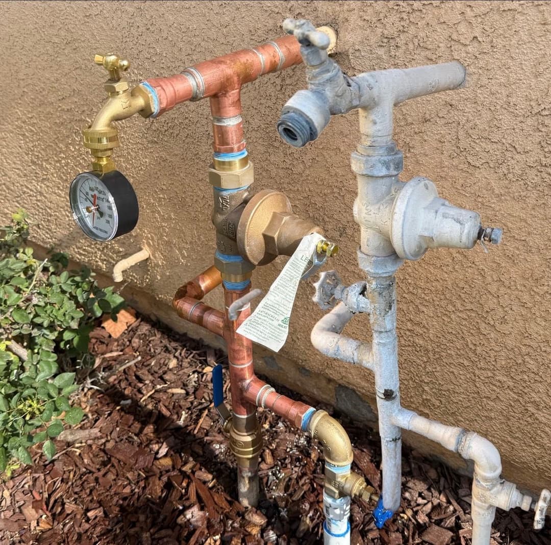 Plumbing system with copper and PVC pipes, pressure gauge, and water valves on exterior wall.