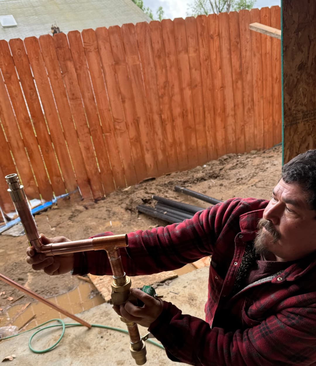 Plumber assembling copper pipes in a construction setting with a wooden fence backdrop.