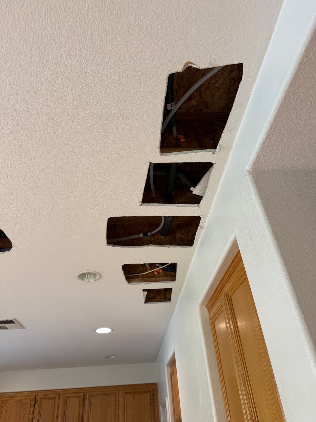 Ceiling with multiple cutouts exposing wiring and pipes in a kitchen renovation.