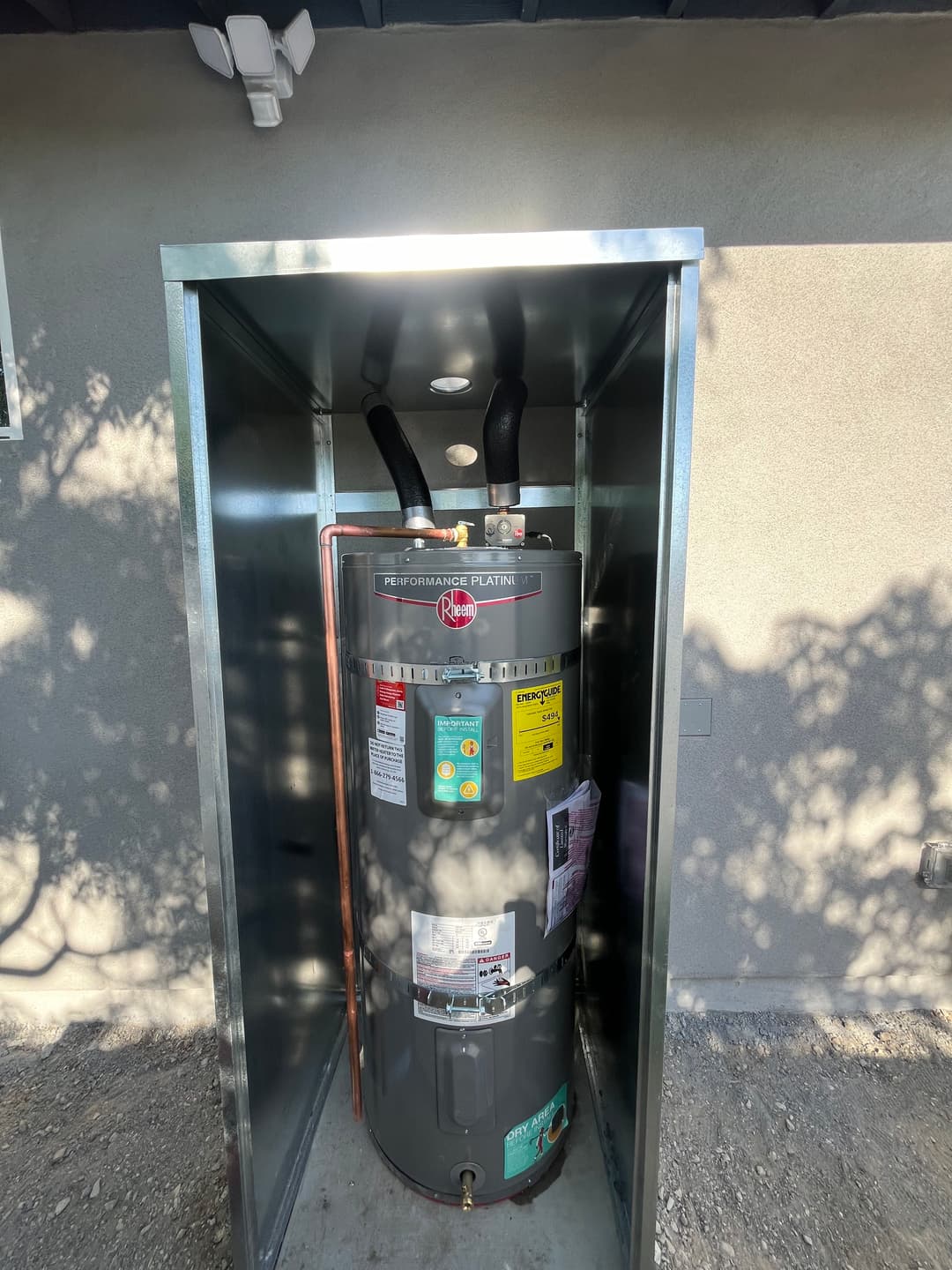Rheem water heater in a protective metal housing with labels and connections visible.