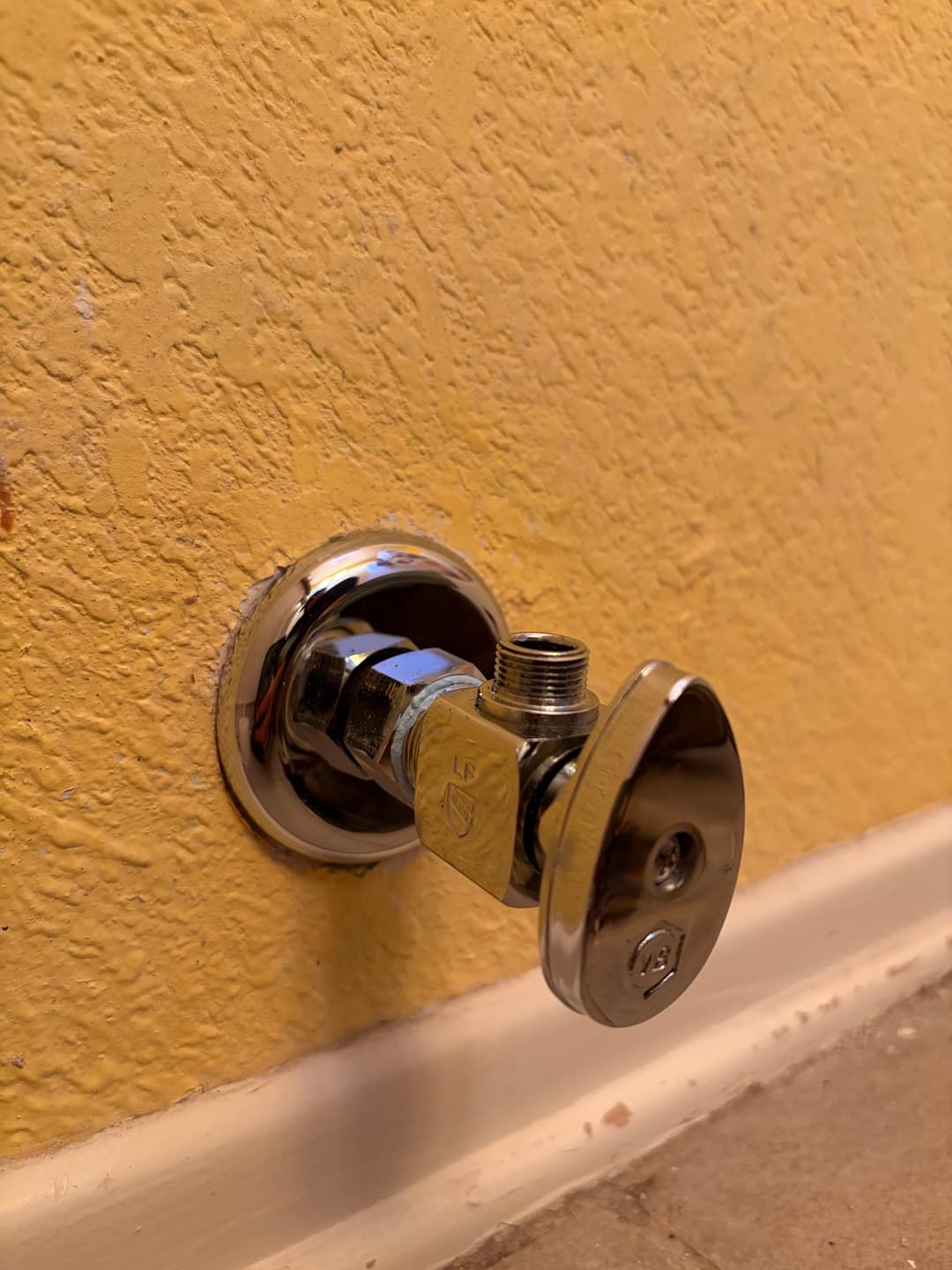 Chrome wall-mounted water shut-off valve on yellow textured wall.