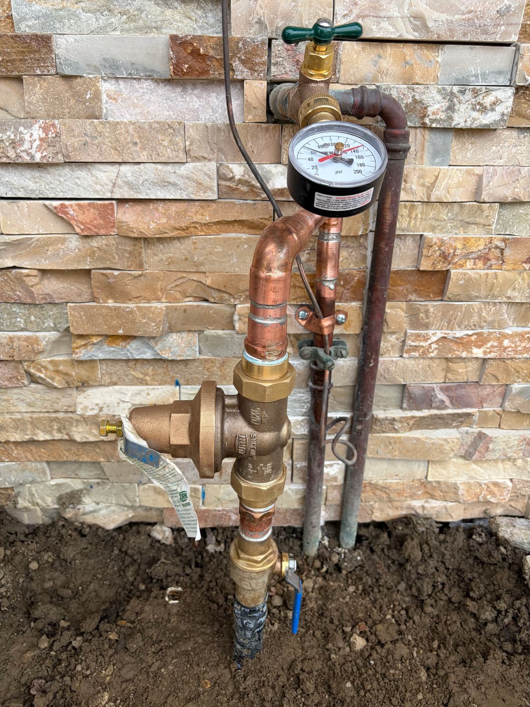 Water pressure regulator with gauge and copper piping against a stone wall backdrop.