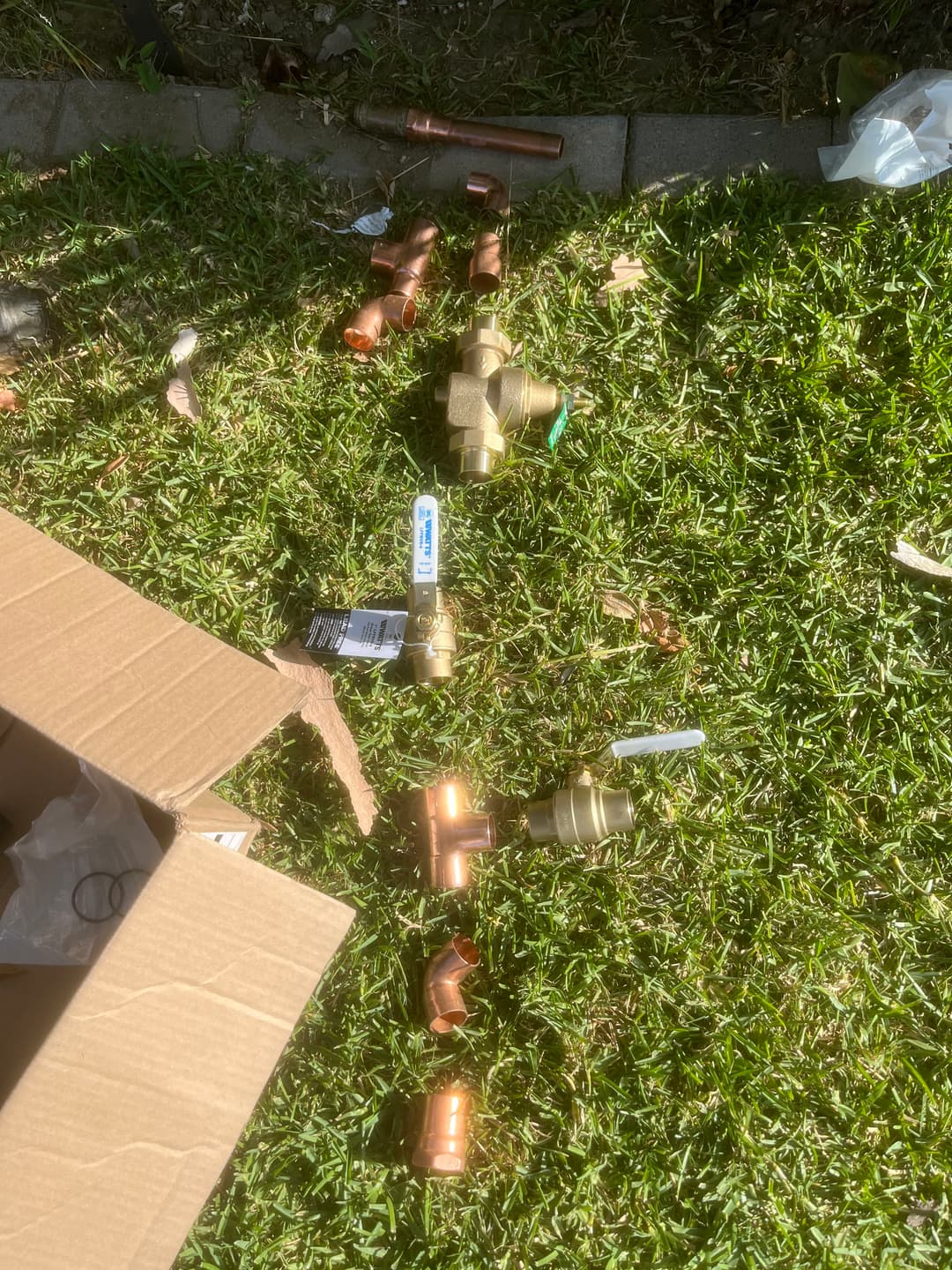 Assorted plumbing fittings and connectors scattered on grass next to an open cardboard box.