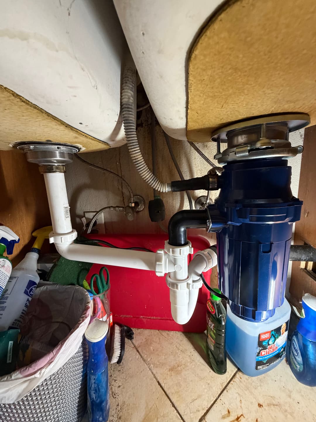 Under-sink plumbing with garbage disposal, pipes, and cleaning supplies in kitchen.