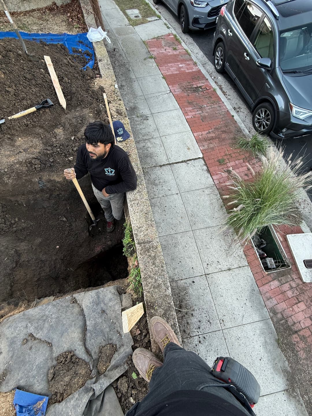 Person digging a trench in a residential area with parked cars nearby.