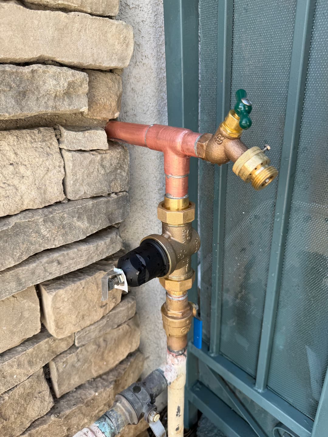 Copper and brass plumbing pipes with a green valve mounted on a stone wall.
