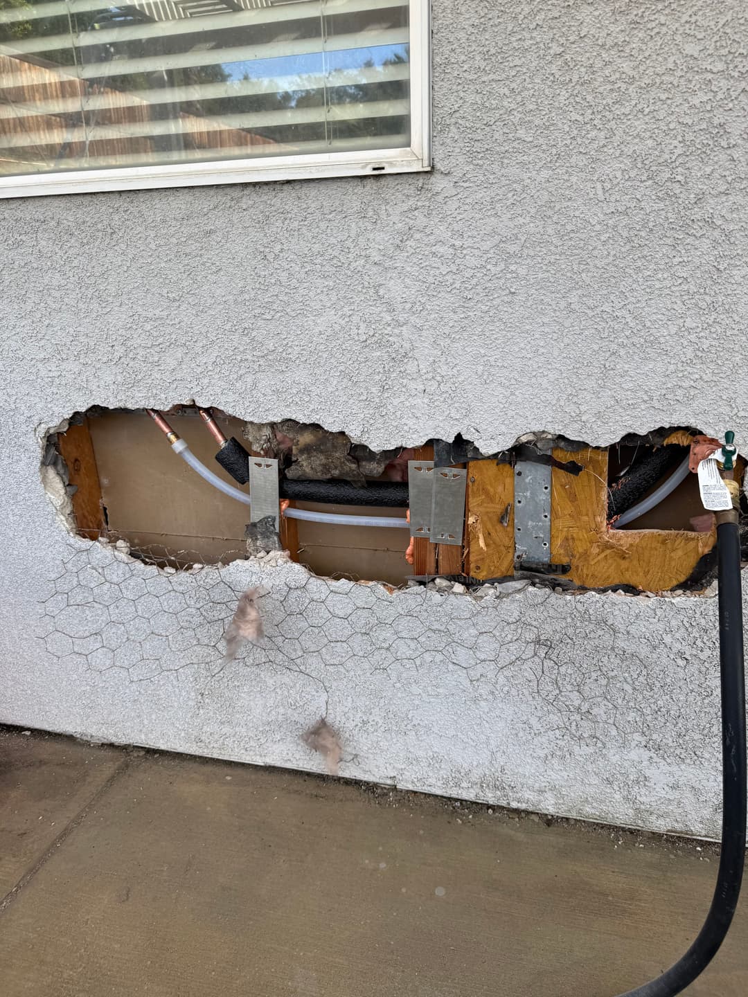 Damaged wall with large hole exposing pipes and insulation, showing structural issues.