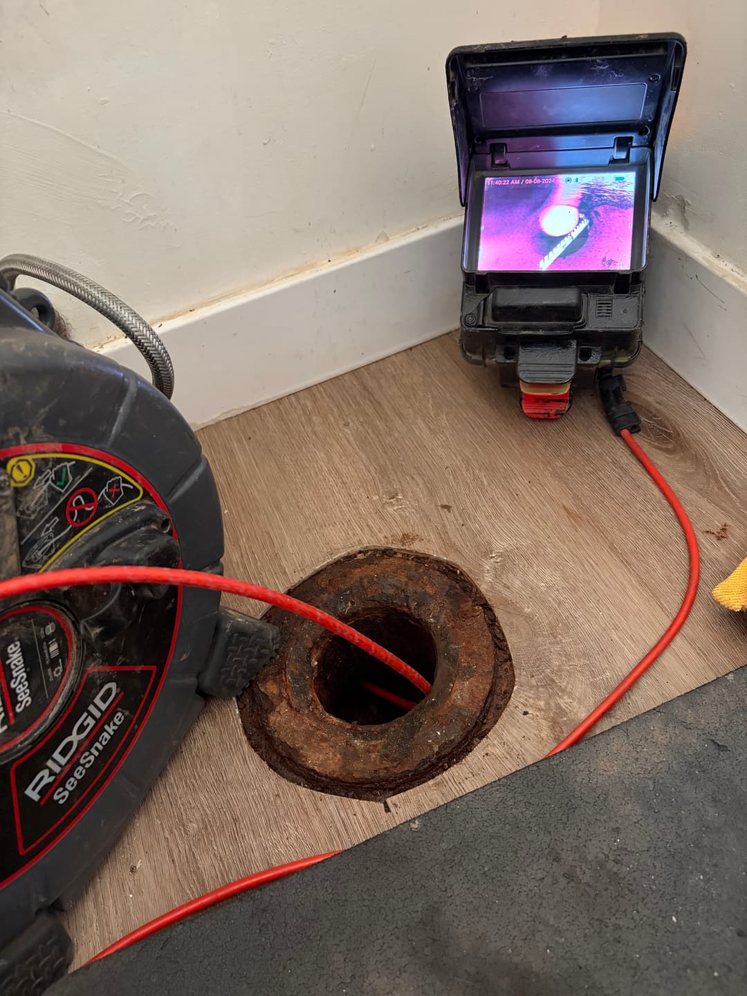 Drain inspection setup with camera monitor and snake tool near rusty pipe opening.