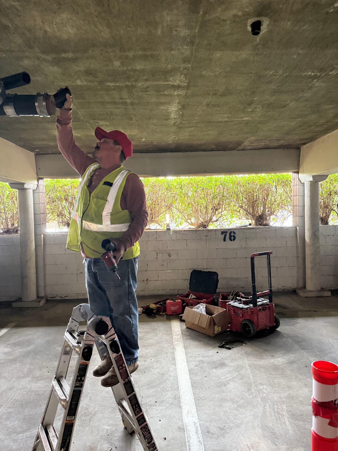 Worker repairing ceiling in a parking garage using tools on a ladder. Tools and equipment visible.