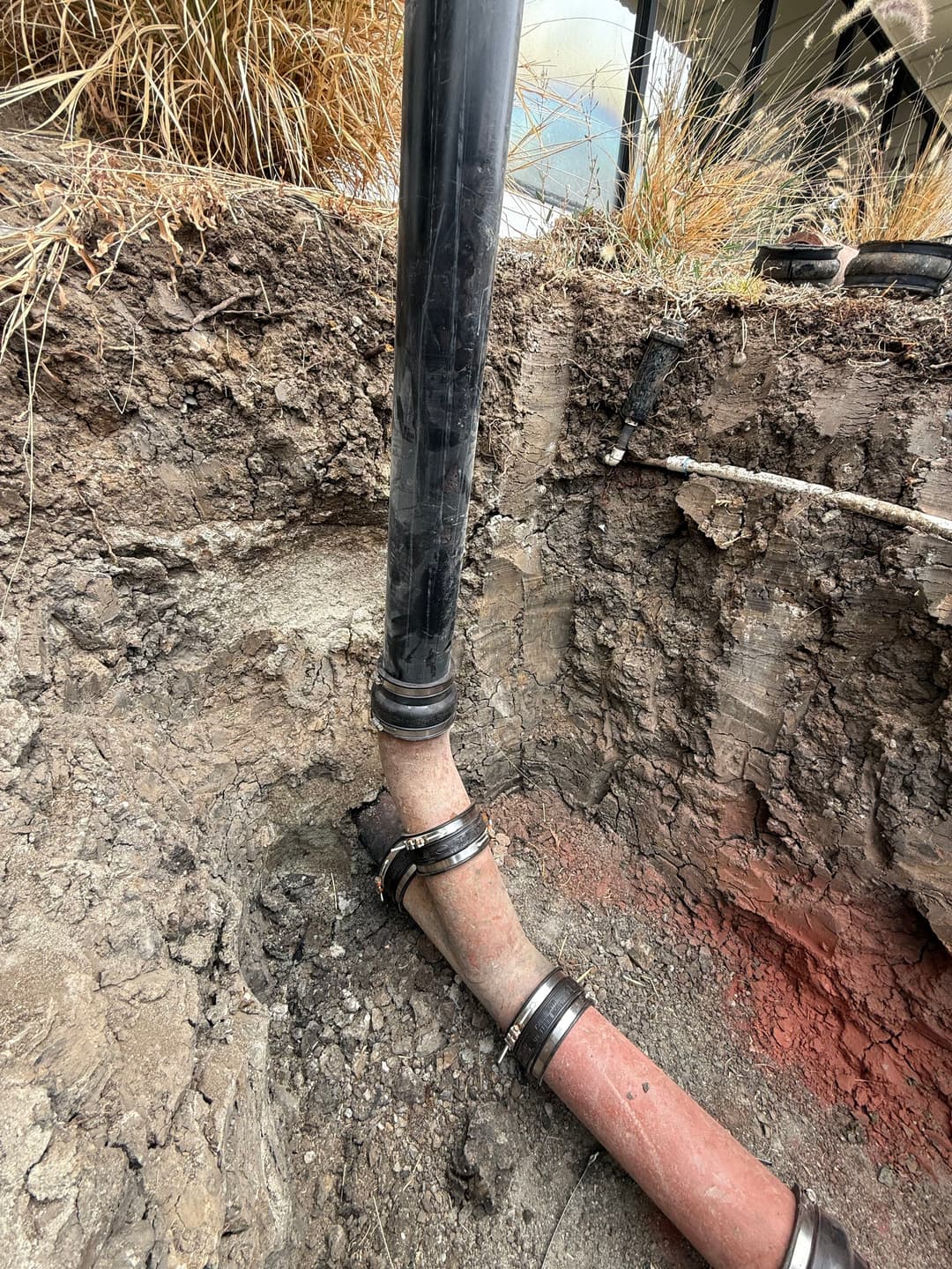Broken underground pipe connected to a black drainage pipe in a dry, excavated area.