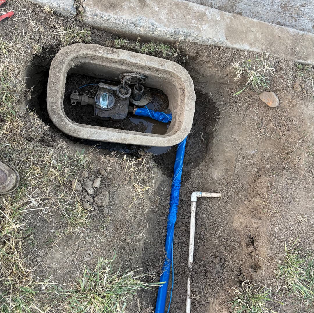 Water meter installation with blue piping in a trench by the sidewalk.