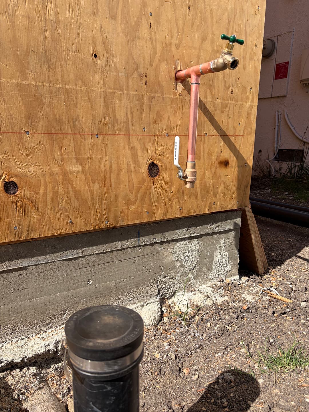 Newly installed exterior water faucet on wood panel wall with concrete base and plumbing.