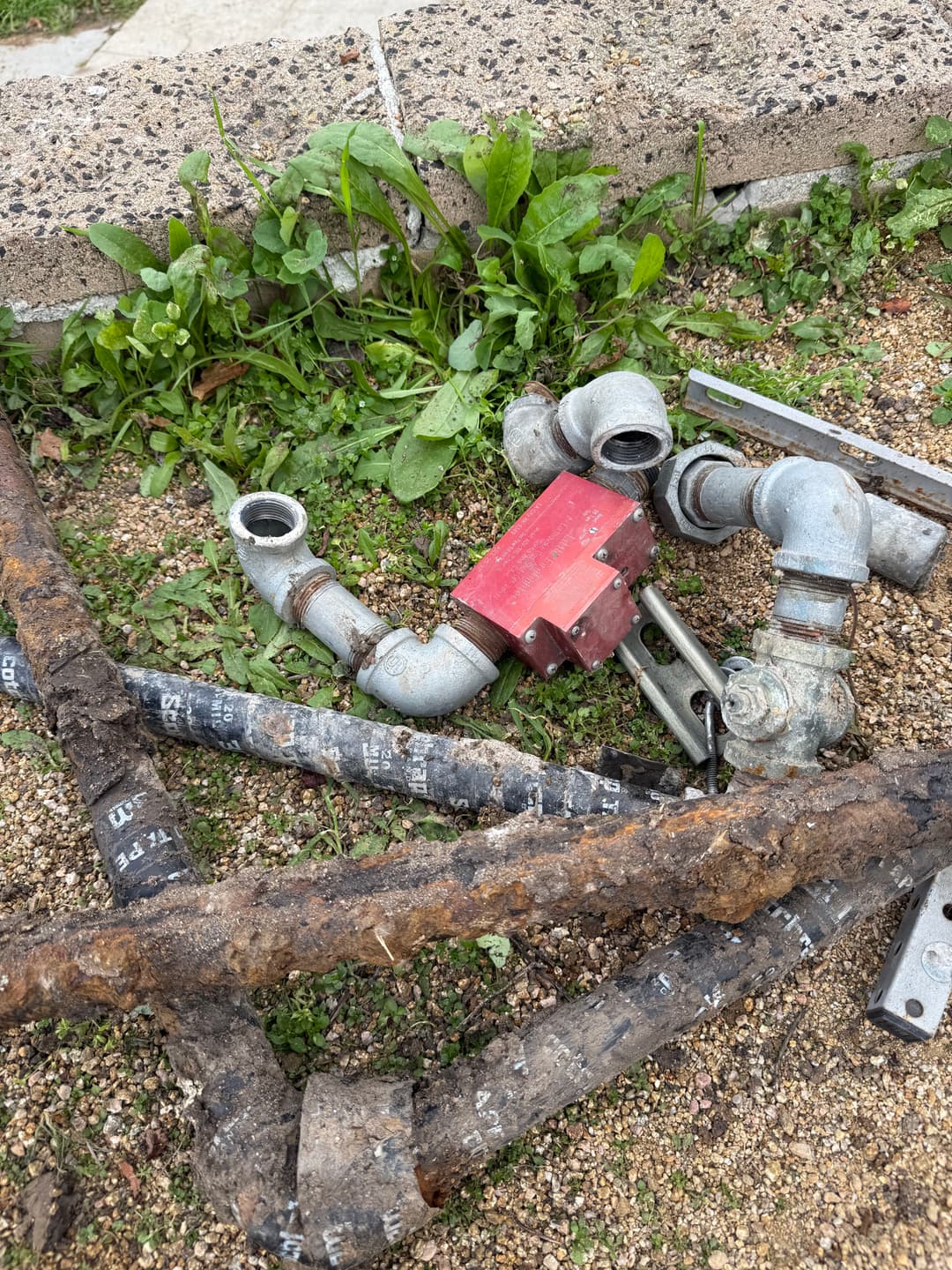 Rusty metal pipes and fittings with a red valve on a gravel surface surrounded by green grass.